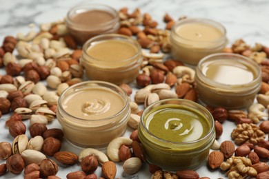 Photo of Jars with butters made of different nuts and ingredients on white marble table, closeup