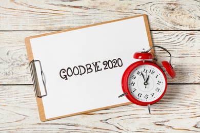 Photo of Clipboard with phrase Goodbye 2020 and alarm clock on white wooden table, flat lay