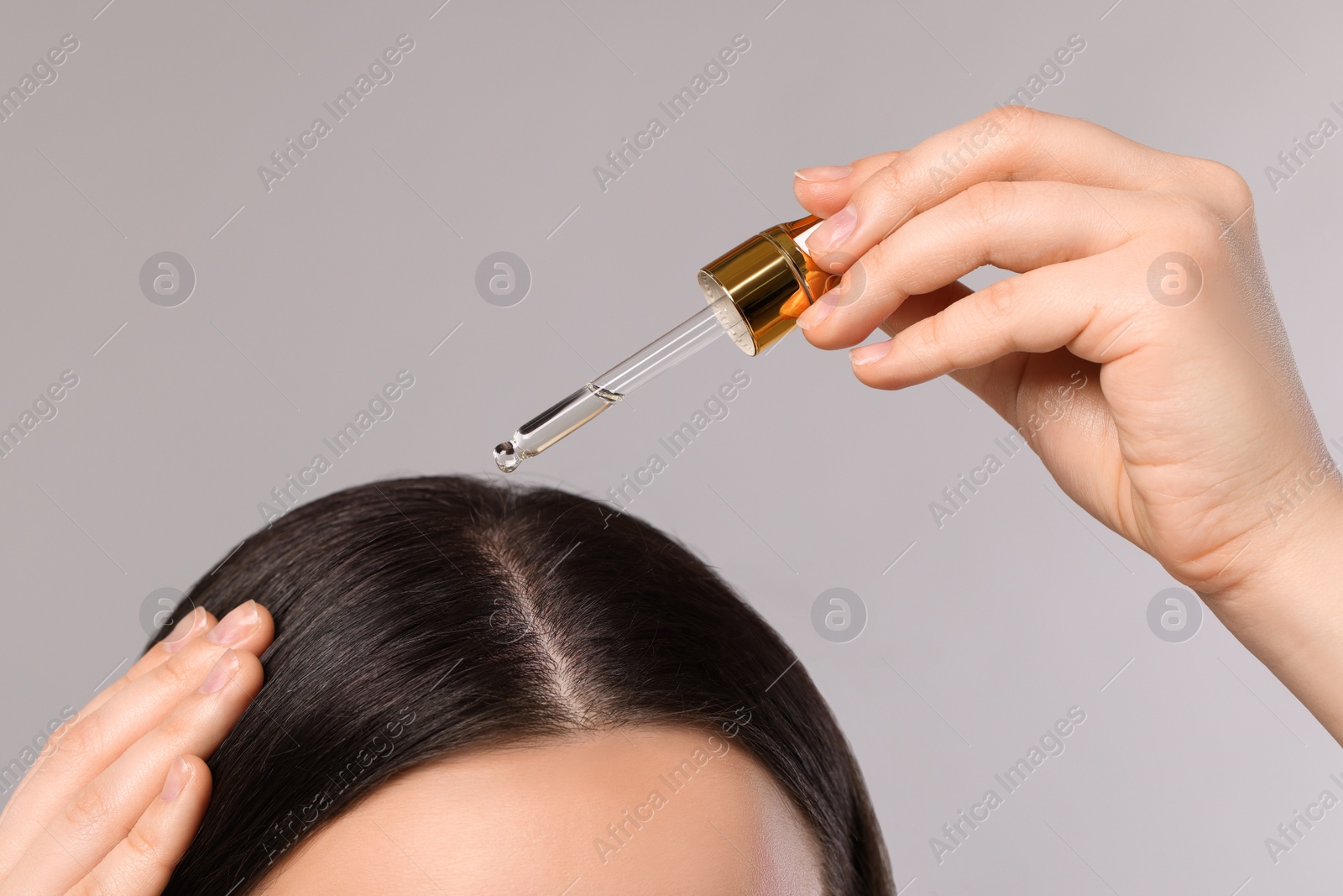 Photo of Woman applying essential oil onto hair roots on light grey background, closeup