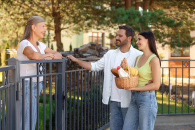 Photo of Friendly relationship with neighbours. Young couple with wicker basket of products treating senior woman near fence outdoors