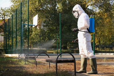 Photo of Person in hazmat suit spraying disinfectant onto bench outdoors. Surface treatment during coronavirus pandemic