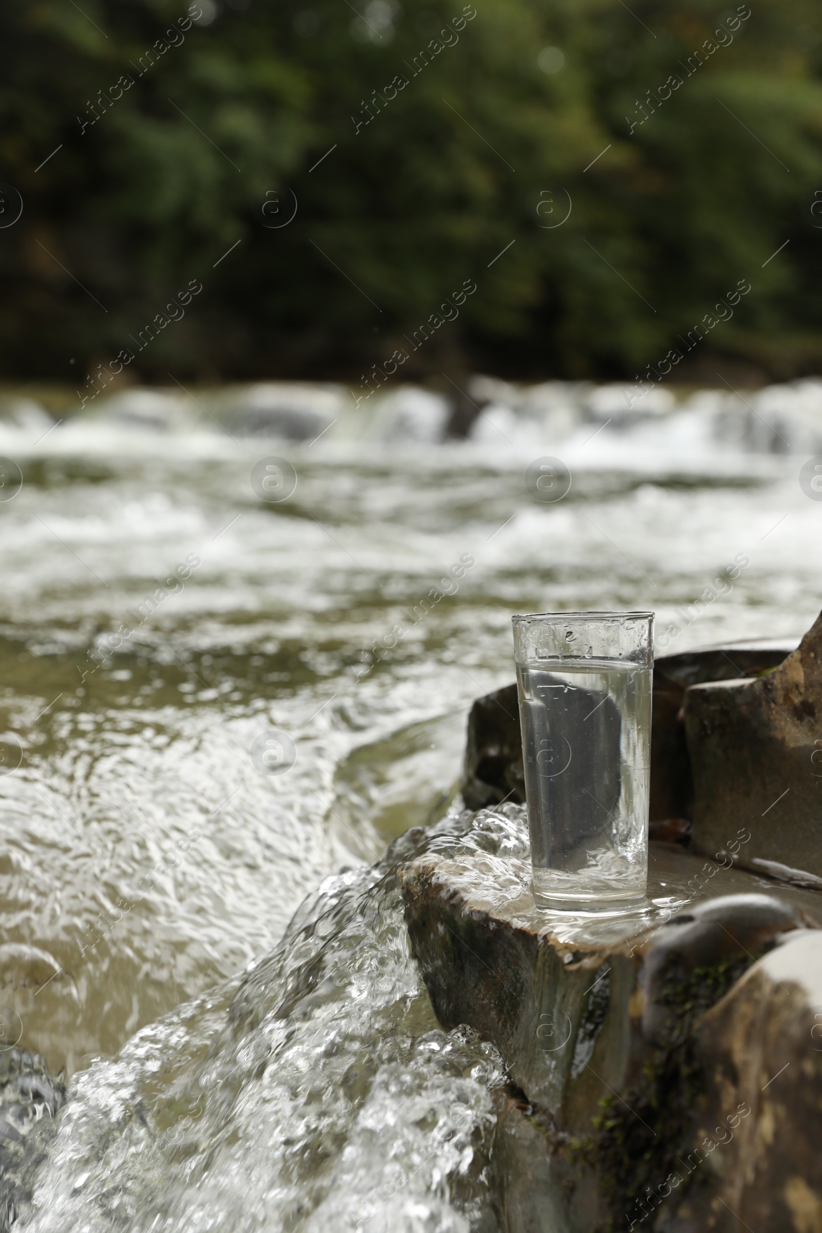 Photo of Glass of fresh water on stone near river, space for text