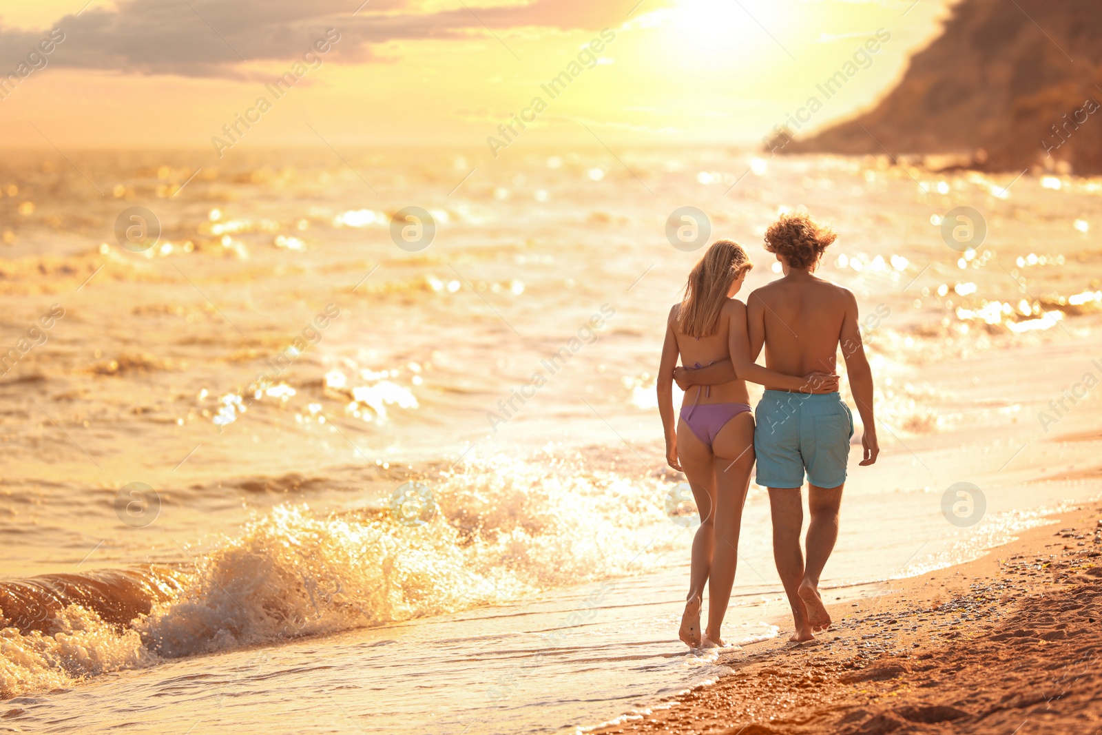 Photo of Young woman in bikini and her boyfriend walking on beach at sunset. Lovely couple