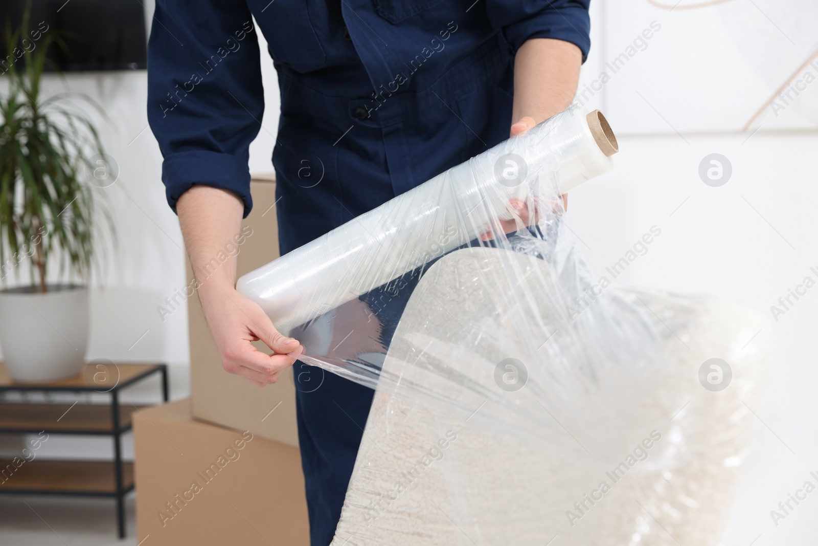 Photo of Male mover with stretch film wrapping armchair in house, closeup