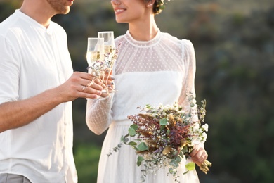 Photo of Happy newlyweds with beautiful field bouquet and glasses of champagne outdoors, closeup