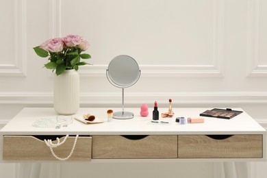 Photo of Mirror, cosmetic products, jewelry and vase with pink roses on white dressing table in makeup room