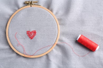 Photo of Embroidered red heart and needle on gray cloth with hoop, top view