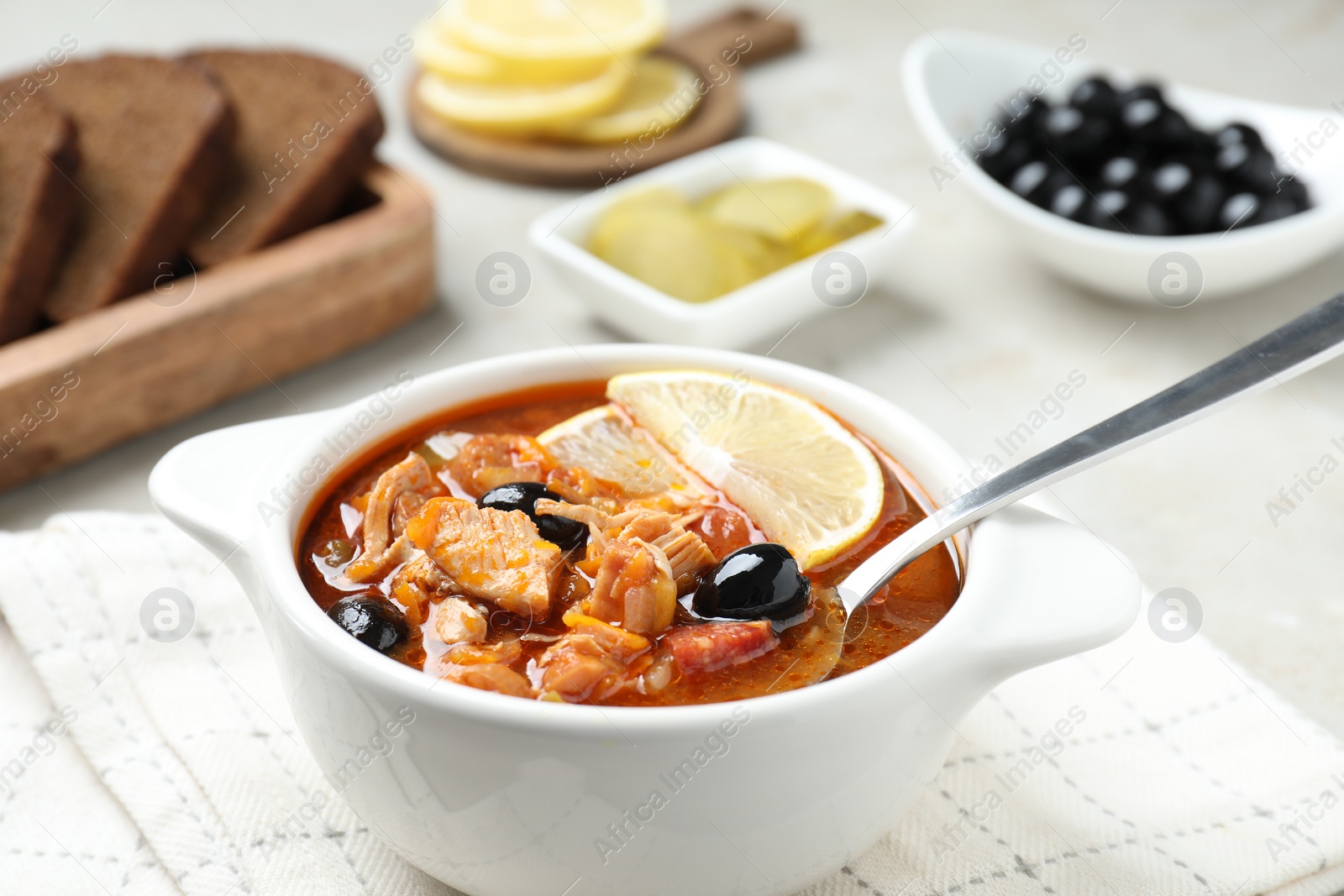 Photo of Meat solyanka soup with sausages, olives, vegetables and spoon in bowl on white table, closeup