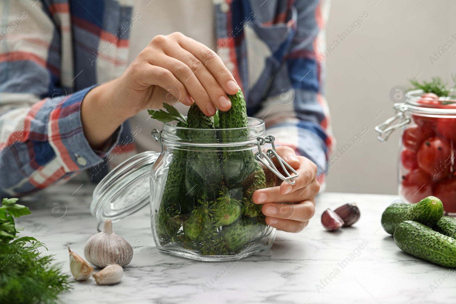 Photo of Woman putting cucumber into glass jar at white marble kitchen table, closeup. Pickling vegetables