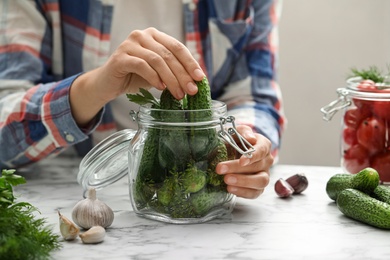 Photo of Woman putting cucumber into glass jar at white marble kitchen table, closeup. Pickling vegetables