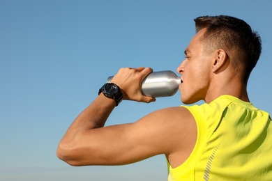 Photo of Young sporty man drinking from water bottle against blue sky on sunny day. Space for text