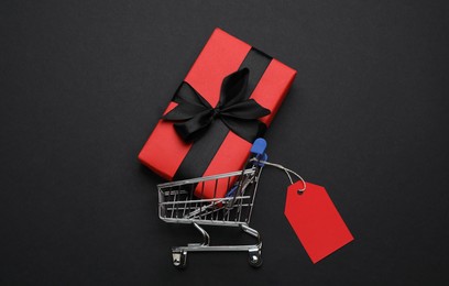 Photo of Shopping cart with price tag and gift box on black background, flat lay
