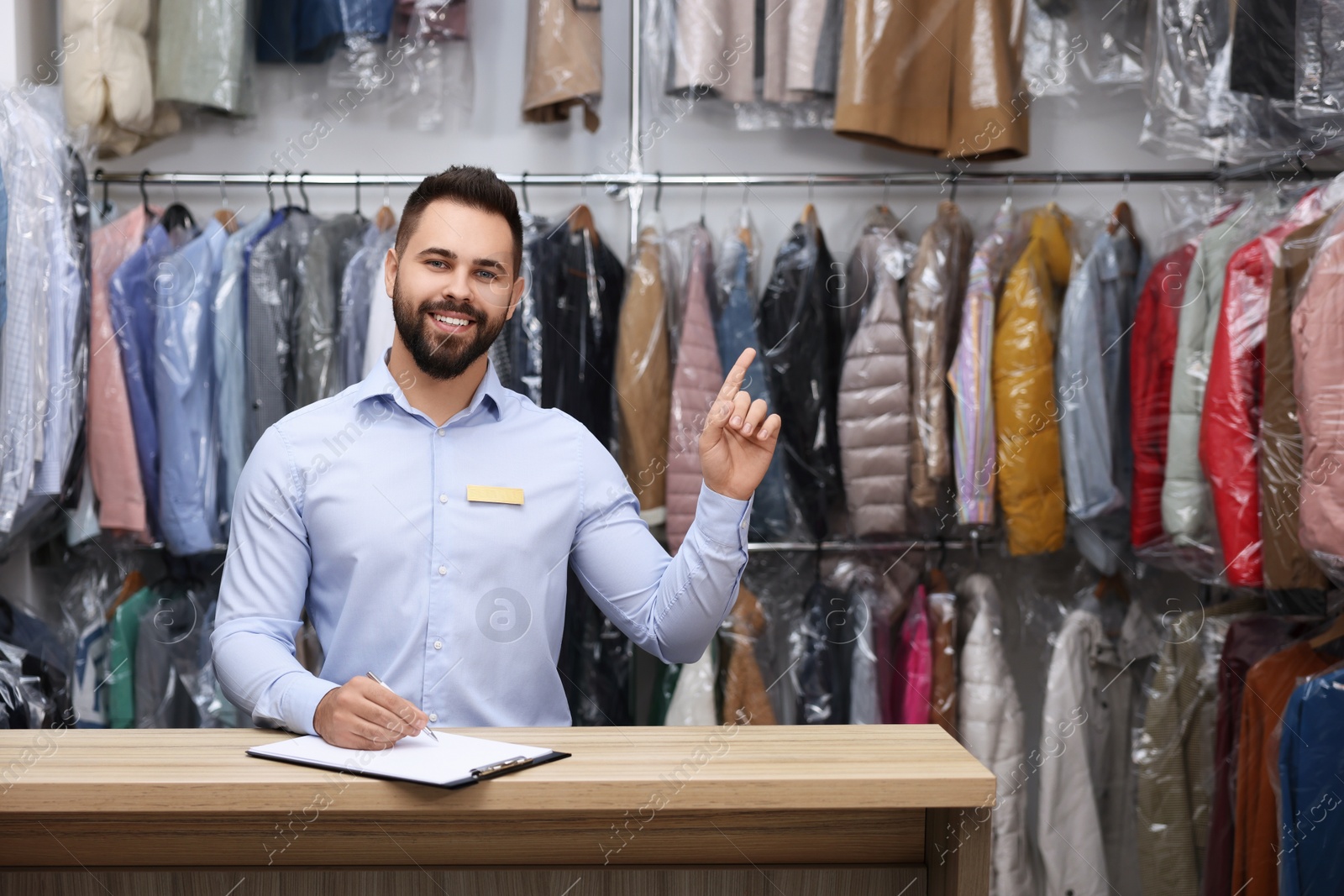 Photo of Dry-cleaning service. Happy worker pointing at something at counter indoors, space for text