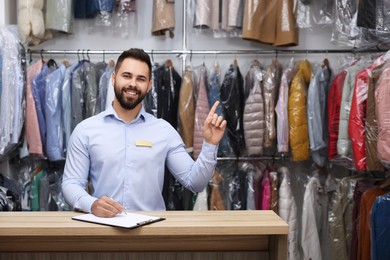 Photo of Dry-cleaning service. Happy worker pointing at something at counter indoors, space for text