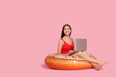 Photo of Young woman using laptop while sitting on inflatable ring against pink background. Space for text