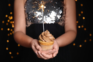 Photo of Woman holding birthday cupcake with sparkler on blurred background, closeup