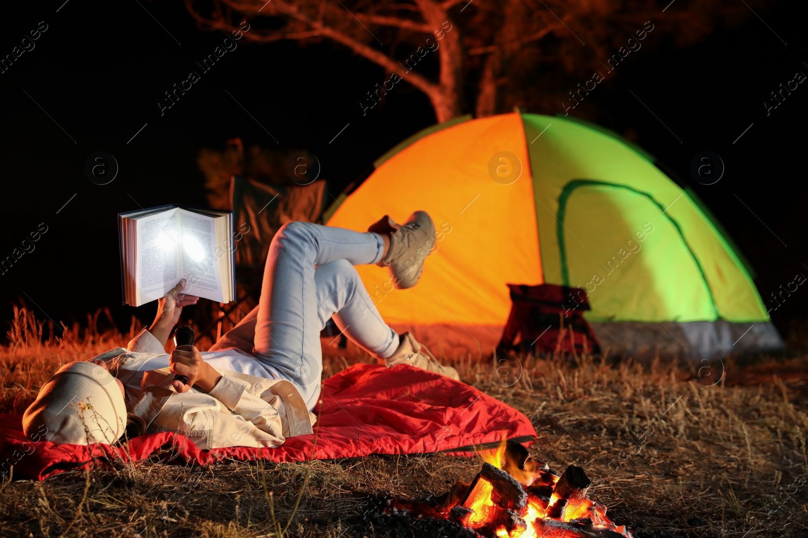 Photo of Young woman with flashlight reading book near bonfire at night. Camping season