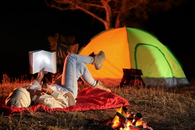 Photo of Young woman with flashlight reading book near bonfire at night. Camping season