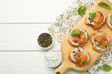 Photo of Tasty canapes with salmon, capers, cucumber and sauce on white wooden table, top view. Space for text