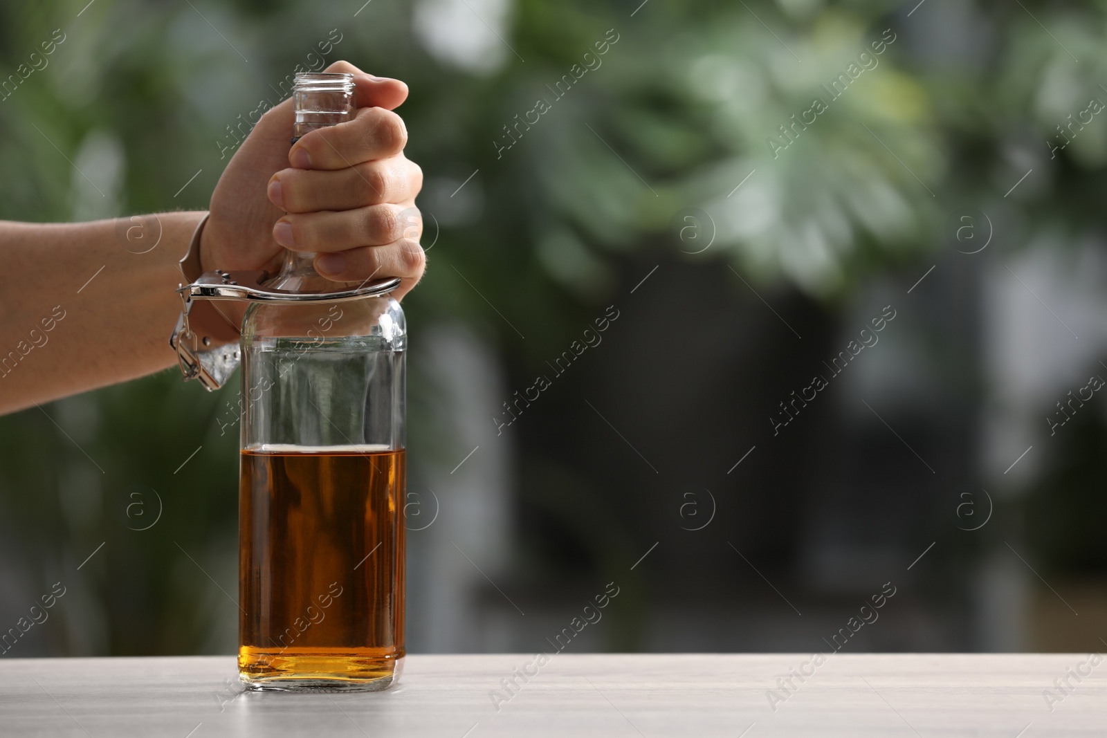 Photo of Addicted man in handcuffs with bottle of alcoholic drink at table against blurred background, closeup. Space for text