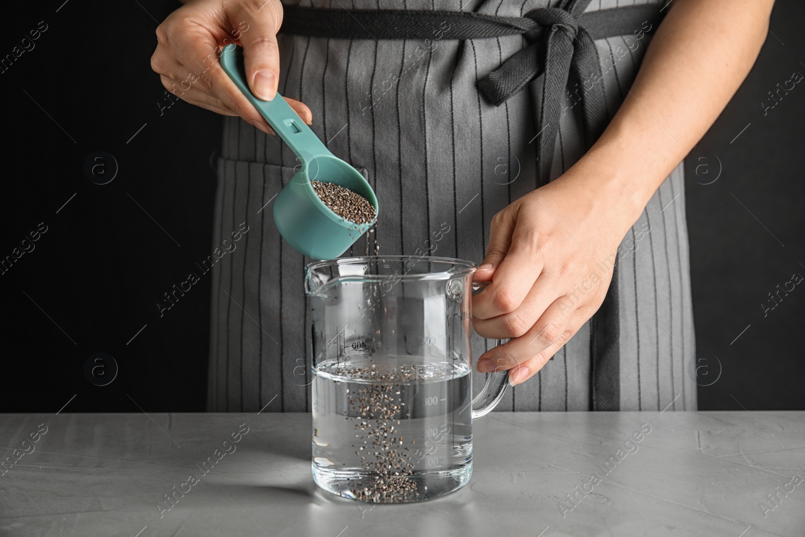 Photo of Young woman pouring chia seeds into measuring glass with water at table, closeup