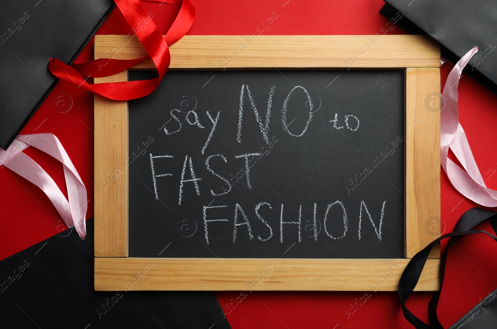 Photo of Small chalkboard with phrase SAY NO TO FAST FASHION and black shopping bags on red background, flat lay