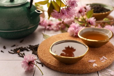 Photo of Traditional ceremony. Cup of brewed tea, teapot, dried leaves and sakura flowers on tiled table, closeup