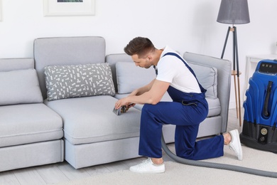 Male worker removing dirt from sofa with professional vacuum cleaner indoors