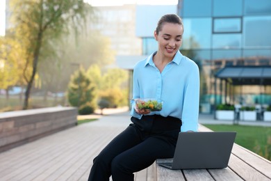 Photo of Smiling businesswoman holding lunch box and working with laptop outdoors. Space for text