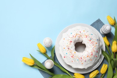 Photo of Delicious Easter cake decorated with sprinkles near beautiful tulips and painted eggs on light blue background, flat lay. Space for text