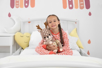 Photo of Cute girl in pajamas with toy deer on bed at home