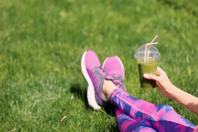 Young woman in sportswear with plastic cup of healthy smoothie on grass outdoors