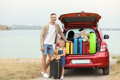 Father and daughter near car trunk with suitcases on riverside. Space for text