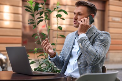 Photo of Handsome man talking on smartphone while using laptop at table in outdoor cafe