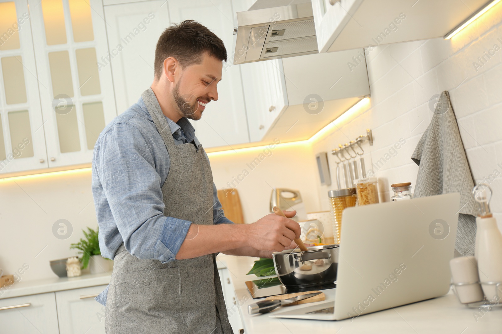 Photo of Man making dinner while watching online cooking course via laptop in kitchen