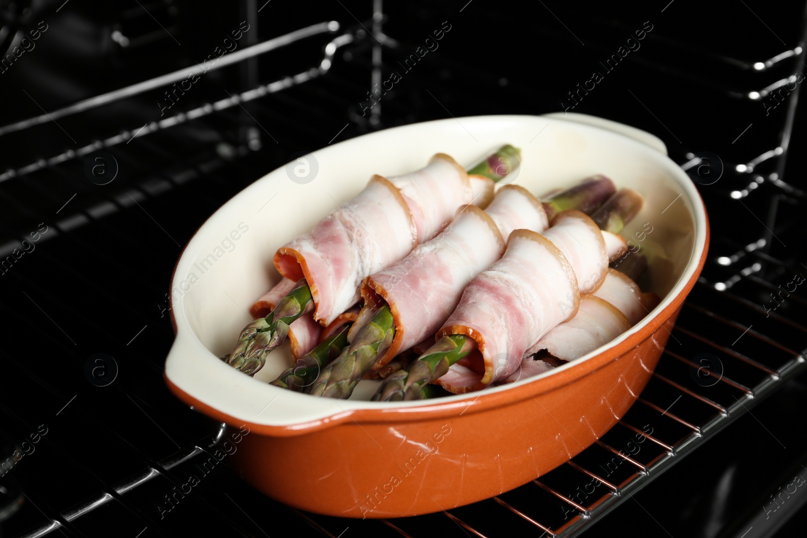 Photo of Uncooked bacon wrapped asparagus in baking dish on oven rack, closeup