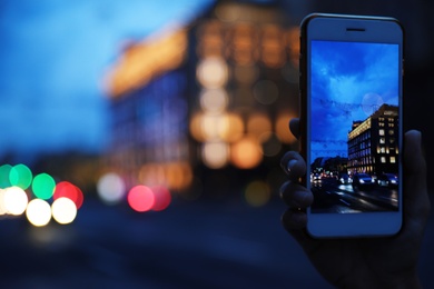 Woman taking photo of modern city in evening with smartphone, focus on hand. Bokeh effect