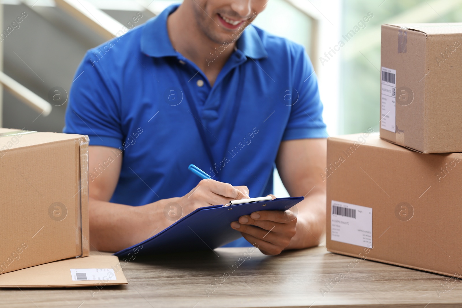 Photo of Young courier working with papers among parcels at table in delivery department