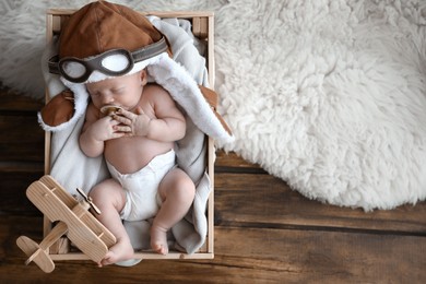 Cute newborn baby wearing aviator hat with toy sleeping in wooden crate, top view. Space for text