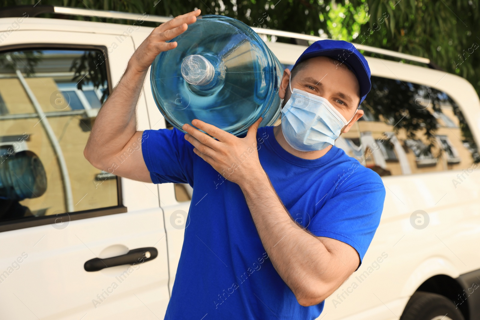 Photo of Courier in medical mask holding bottle of cooler water near car outdoors. Delivery during coronavirus quarantine