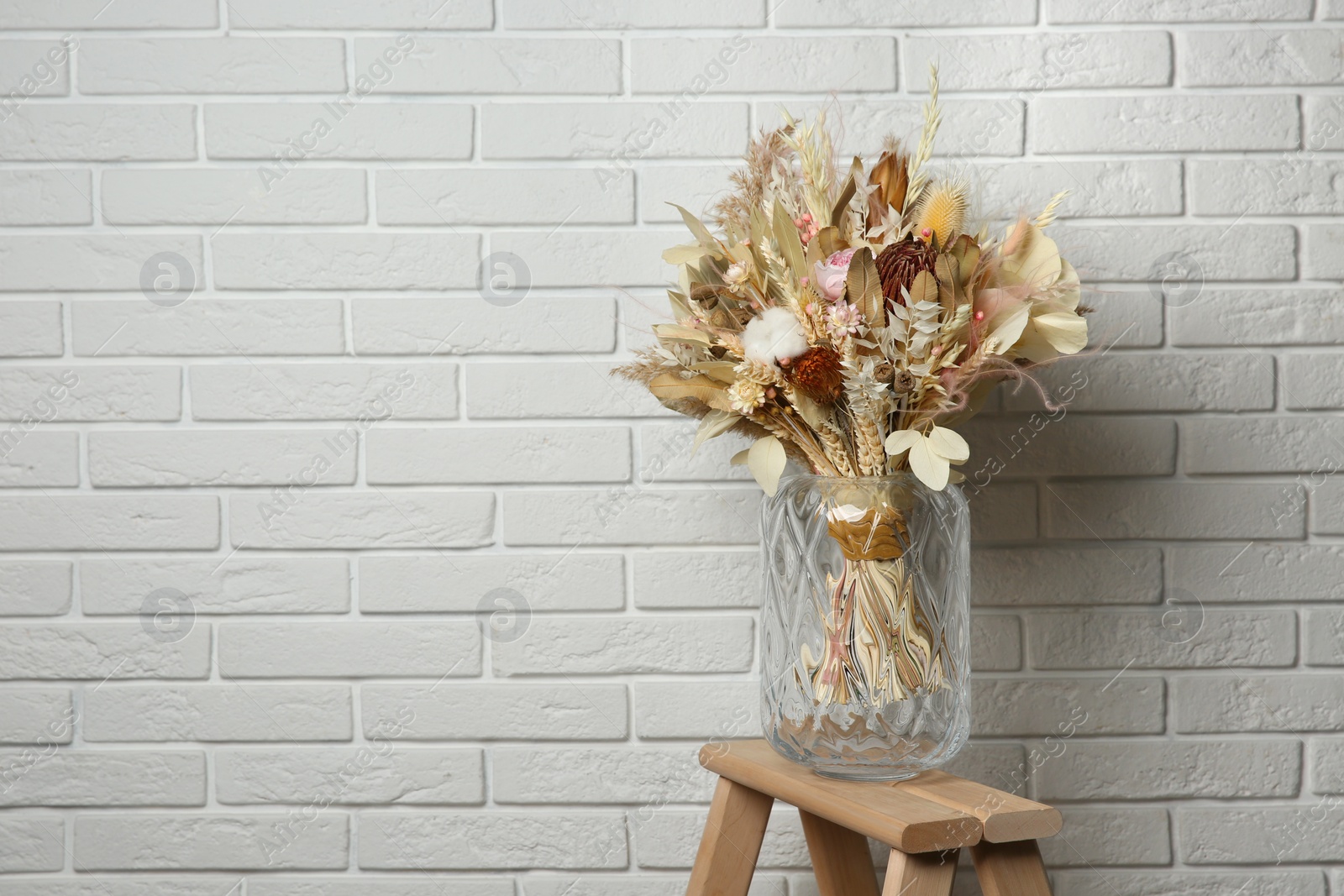 Photo of Beautiful dried flower bouquet in glass vase on wooden ladder near white brick wall. Space for text