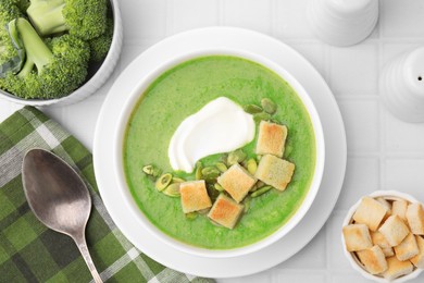 Delicious broccoli cream soup with croutons, sour cream and pumpkin seeds served on white tiled table, flat lay