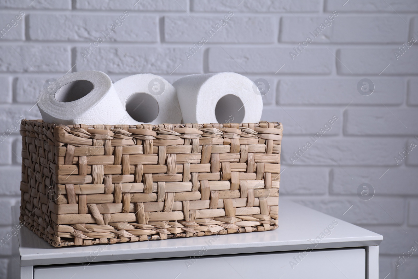 Photo of Toilet paper rolls in wicker basket on chest of drawers near white brick wall