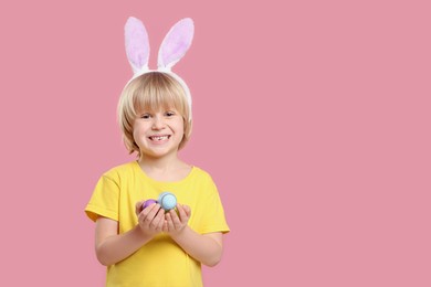 Photo of Happy boy in bunny ears headband holding painted Easter eggs on pink background. Space for text