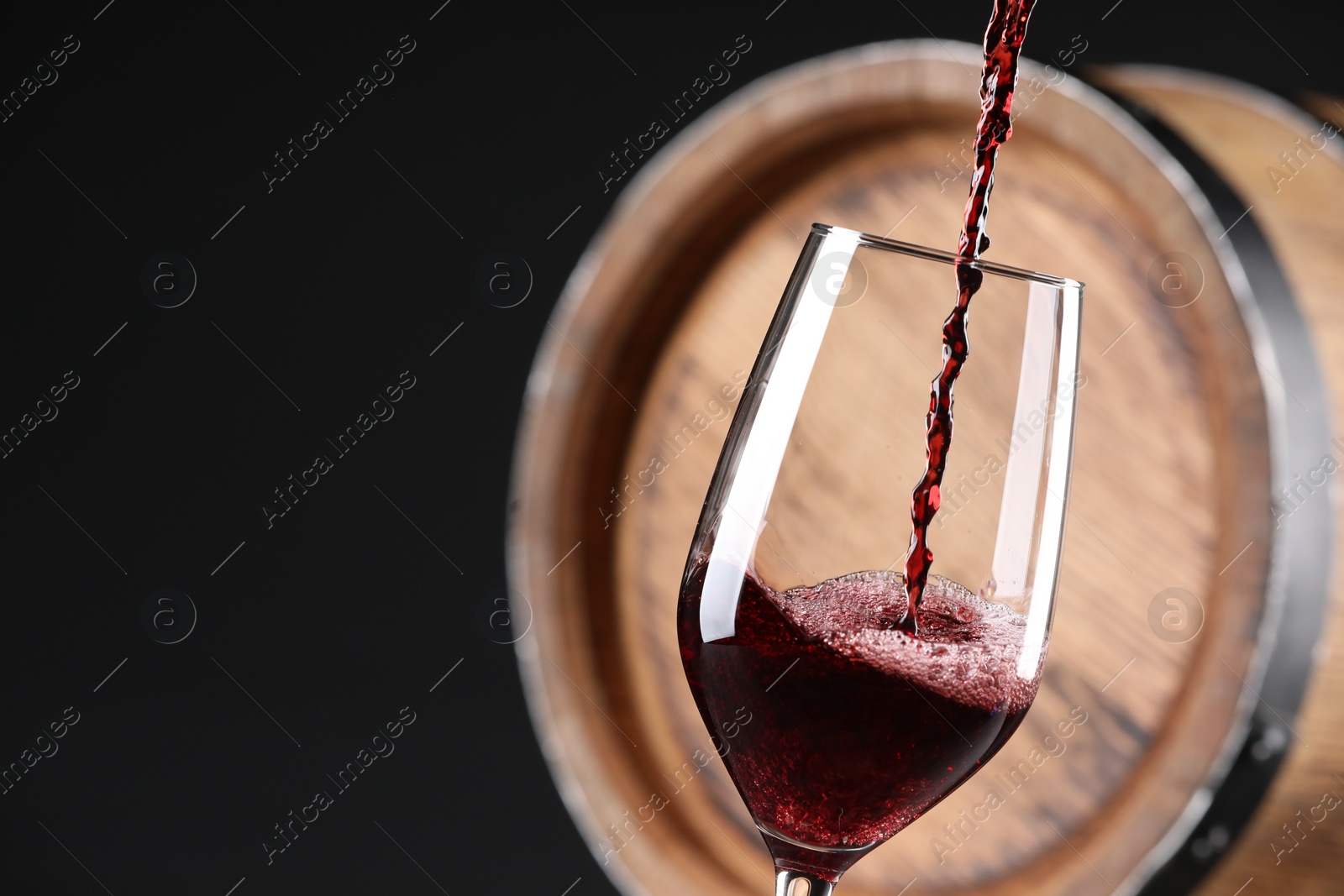 Photo of Pouring red wine into glass near wooden barrel against black background, closeup. Space for text