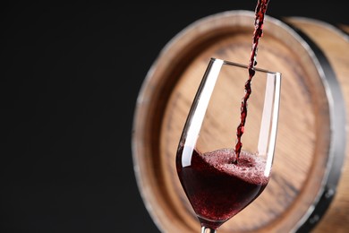 Pouring red wine into glass near wooden barrel against black background, closeup. Space for text