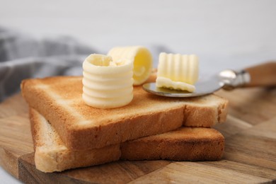 Photo of Tasty butter curls, knife and toasts on wooden board, closeup