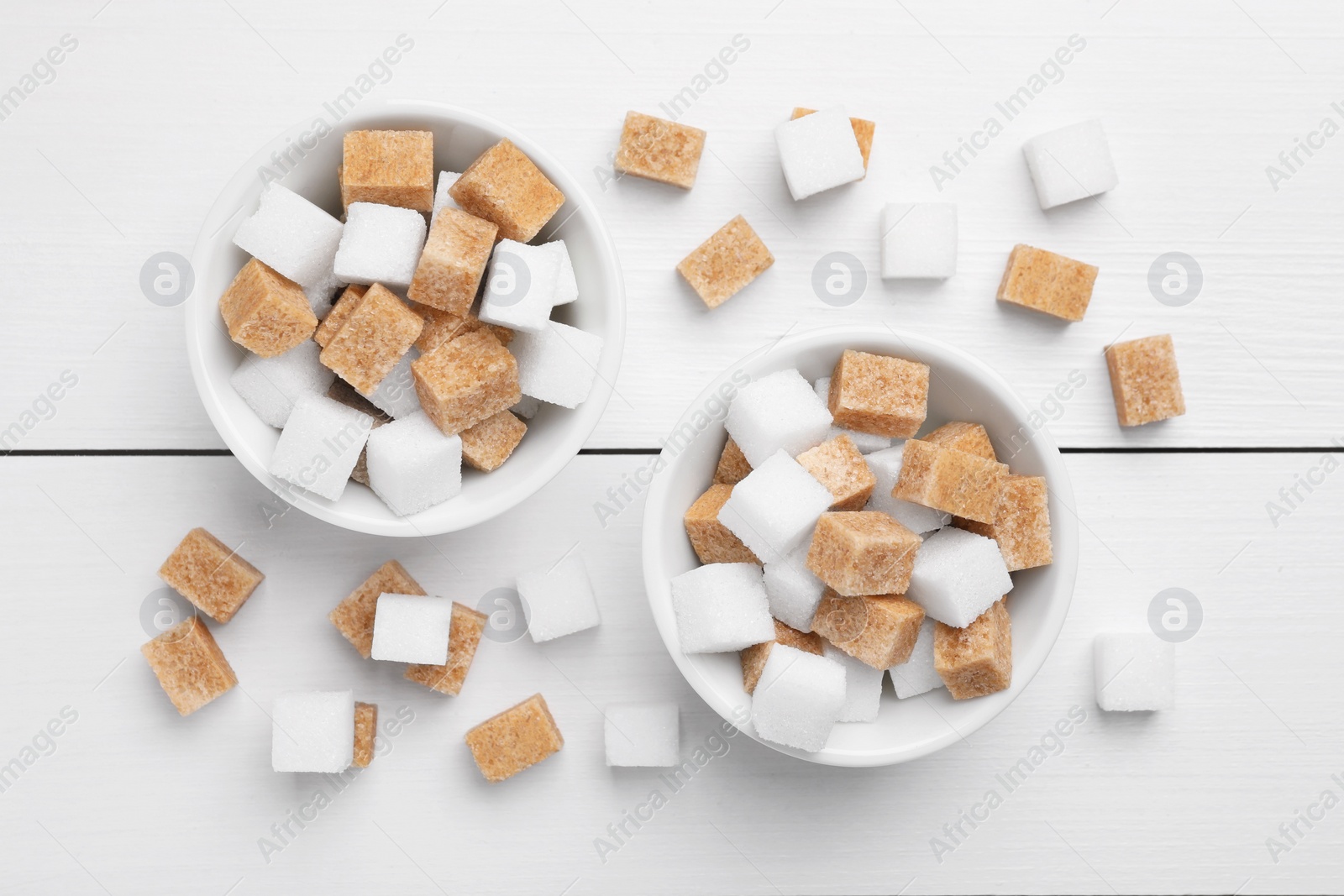 Photo of Different sugar cubes in bowls on white wooden table, flat lay