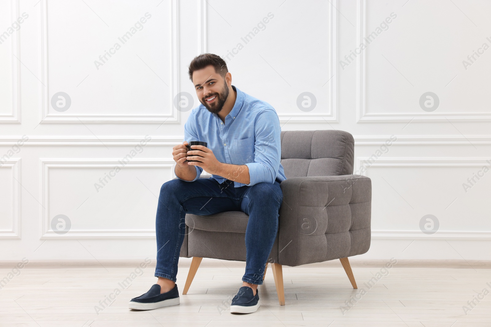 Photo of Handsome man with cup of drink sitting in armchair near white wall indoors