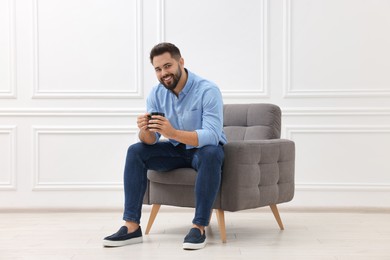 Handsome man with cup of drink sitting in armchair near white wall indoors